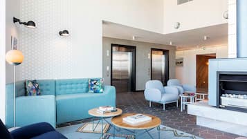 Un vestíbulo moderno con un sofá azul, dos mesas redondas para café, una chimenea y una zona de recepción con ascensor.