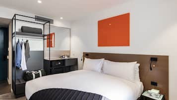 Una habitación de hotel moderna con una cama grande, un armario empotrado, una mesita de noche con lámpara y una obra de arte naranja de gran tamaño en la pared.