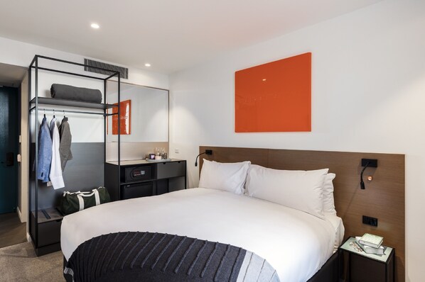 A modern hotel room with a large bed, a built-in wardrobe, a bedside table with a lamp, and a large orange artwork on the wall.
