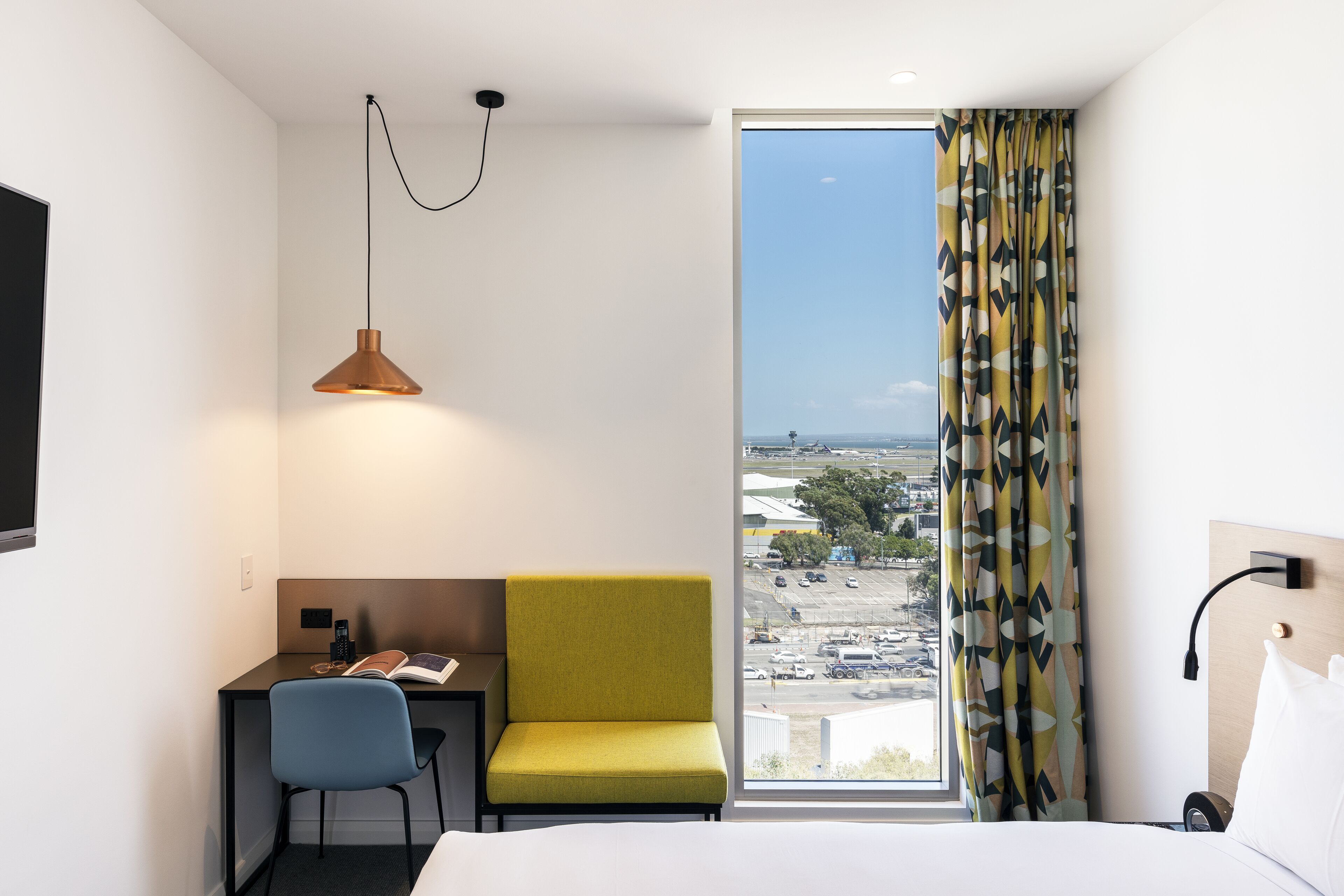 A hotel room with a bed, a desk with a chair, a small table with a book, a lamp, and a view of the city through a large window.