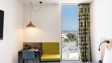 A hotel room with a bed, a desk with a chair, a small table with a book, a lamp, and a view of the city through a large window.