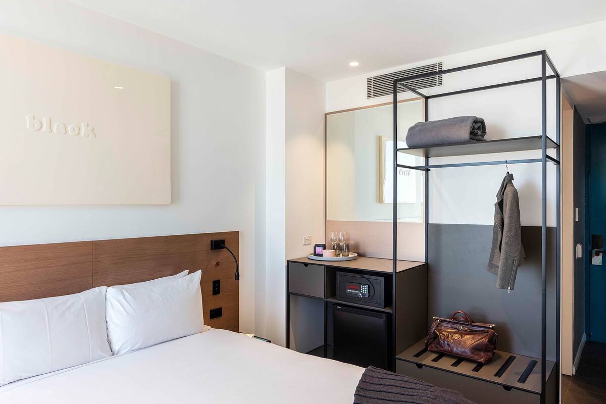 A hotel room with a bed, a desk with a computer, a mirror, a coat rack, and a small safe.