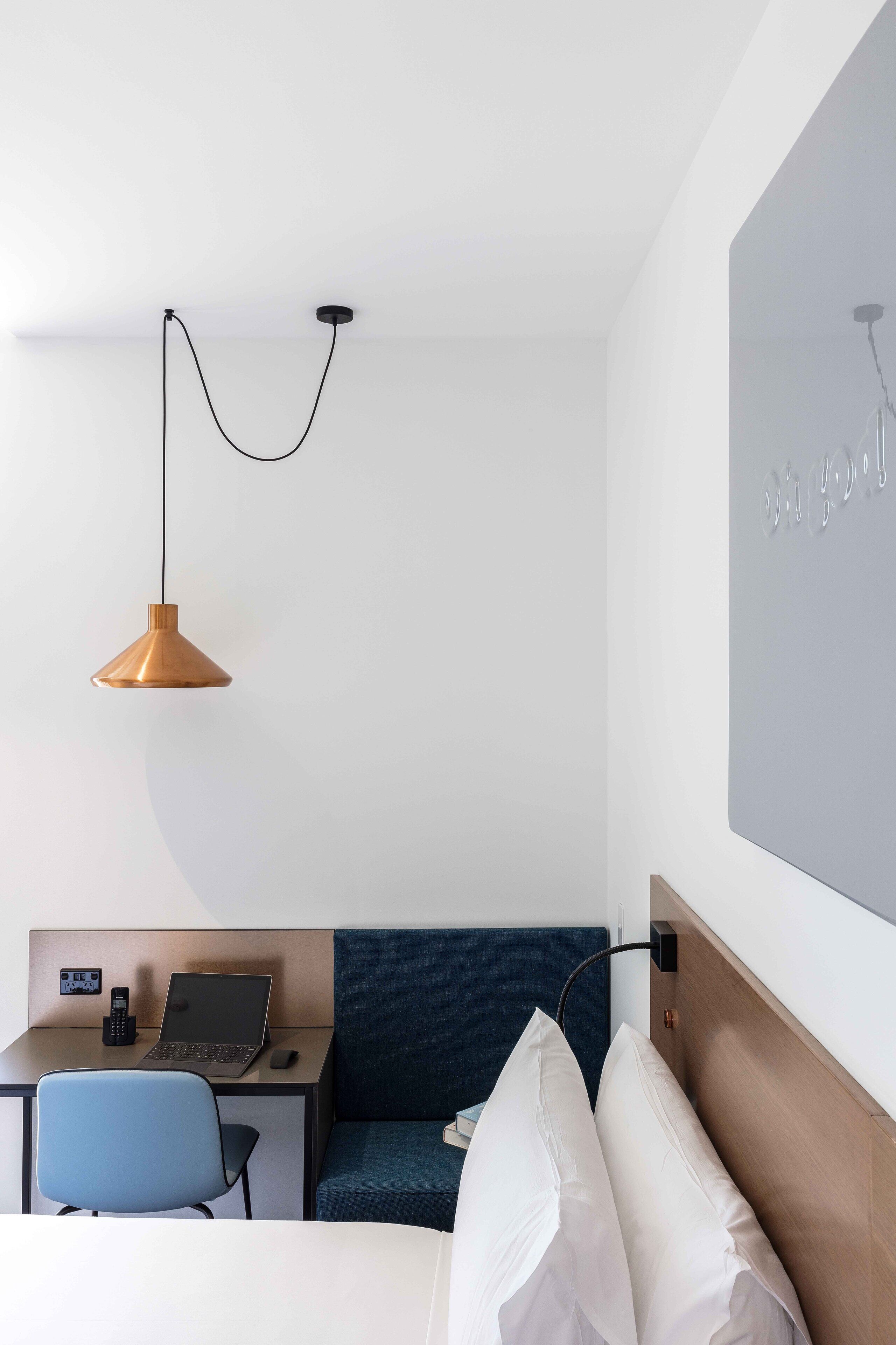 Habitación de hotel con una cama, un escritorio con una computadora portátil, una silla, un sofá azul y una lámpara colgante.