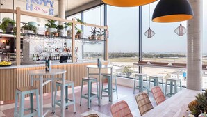 Zona de bar con un letrero de 'HAPPY HOUR', mostrador de bar con botellas y asientos con vista al aeropuerto.
