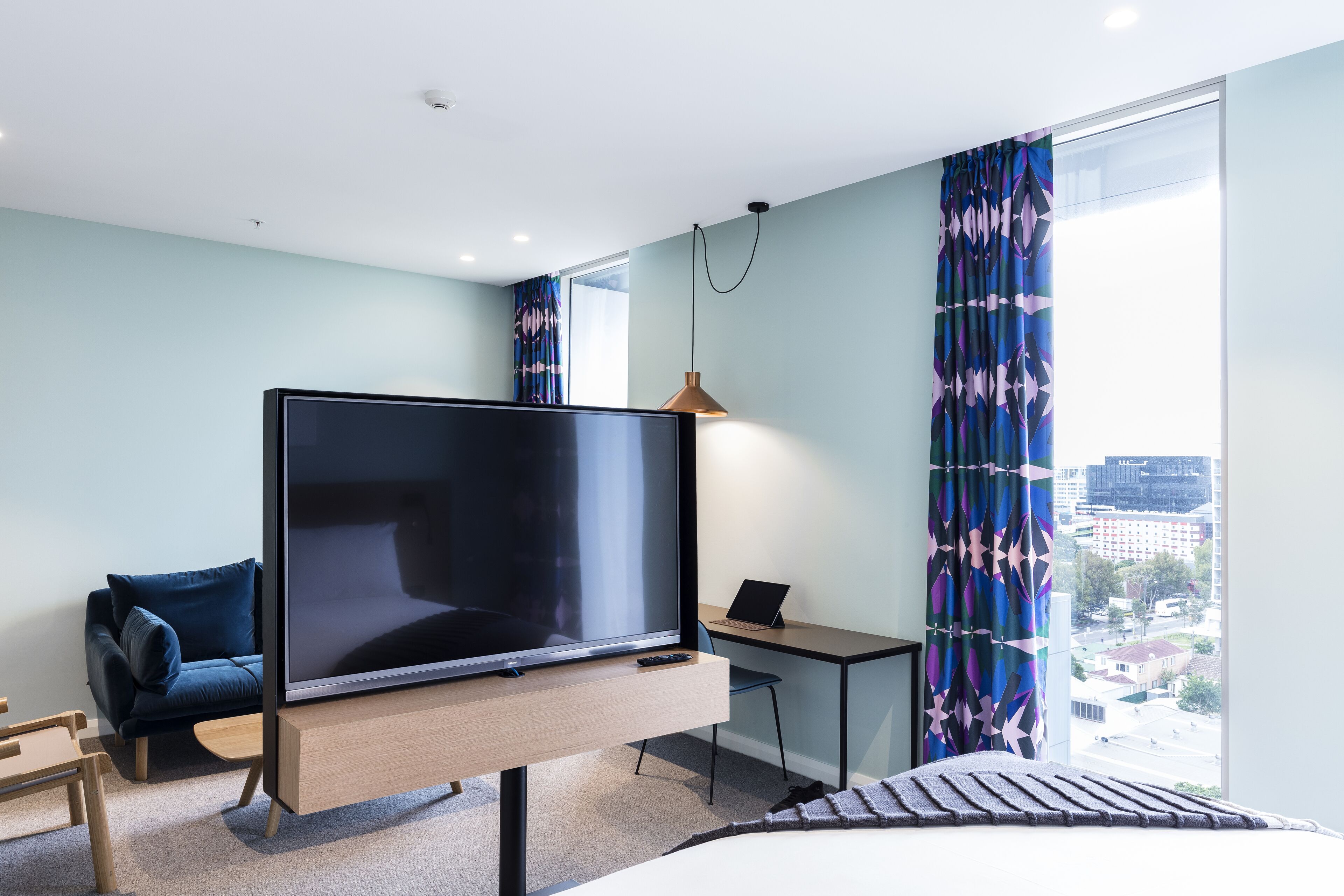 Una habitación de hotel moderna con un televisor de pantalla plana grande, un escritorio con una computadora portátil, un sofá cómodo y un ventanal con vistas a la ciudad.