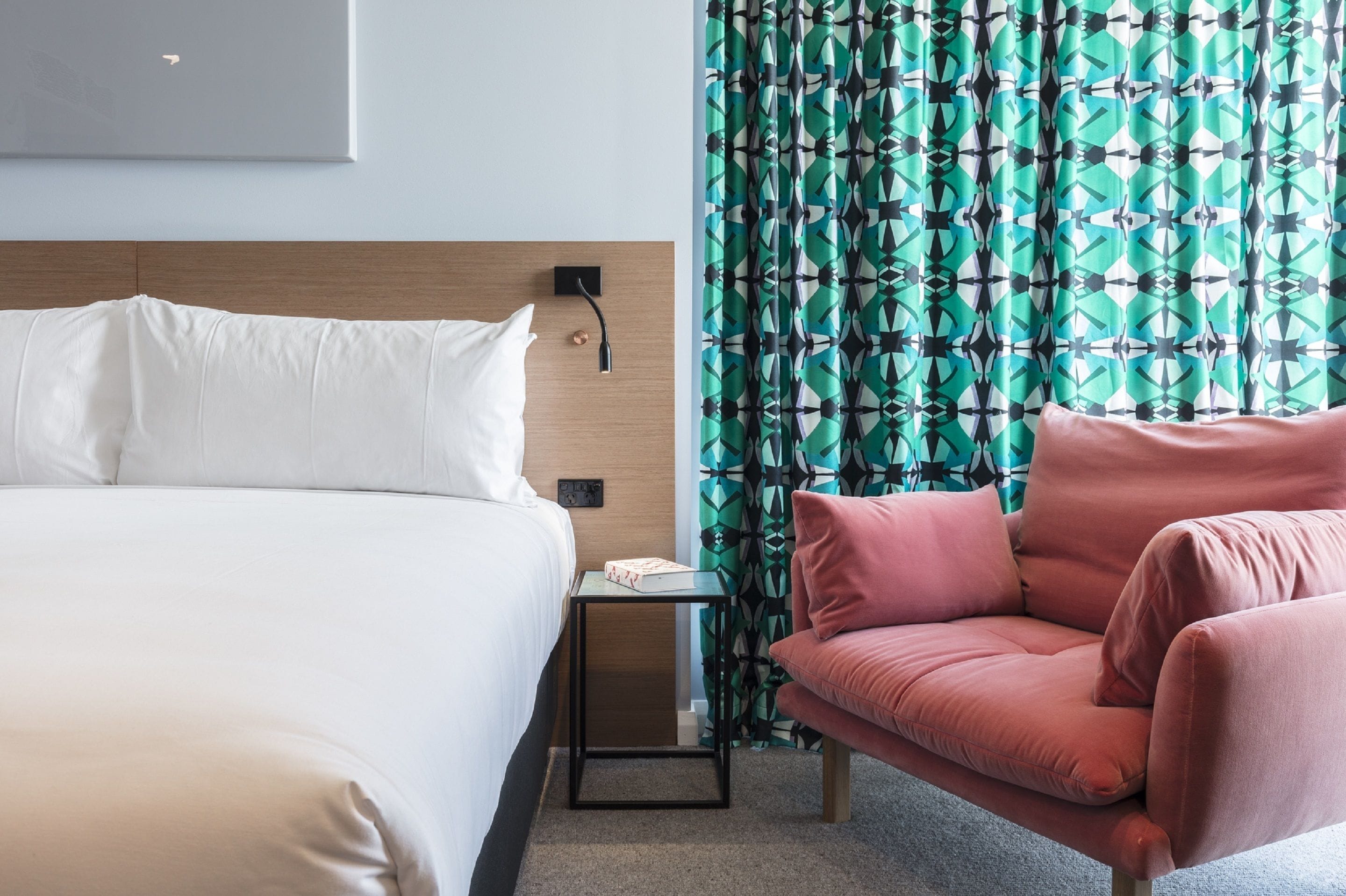 Une chambre d’hôtel avec un lit, une petite table de chevet, une chaise rose et des rideaux à motifs.