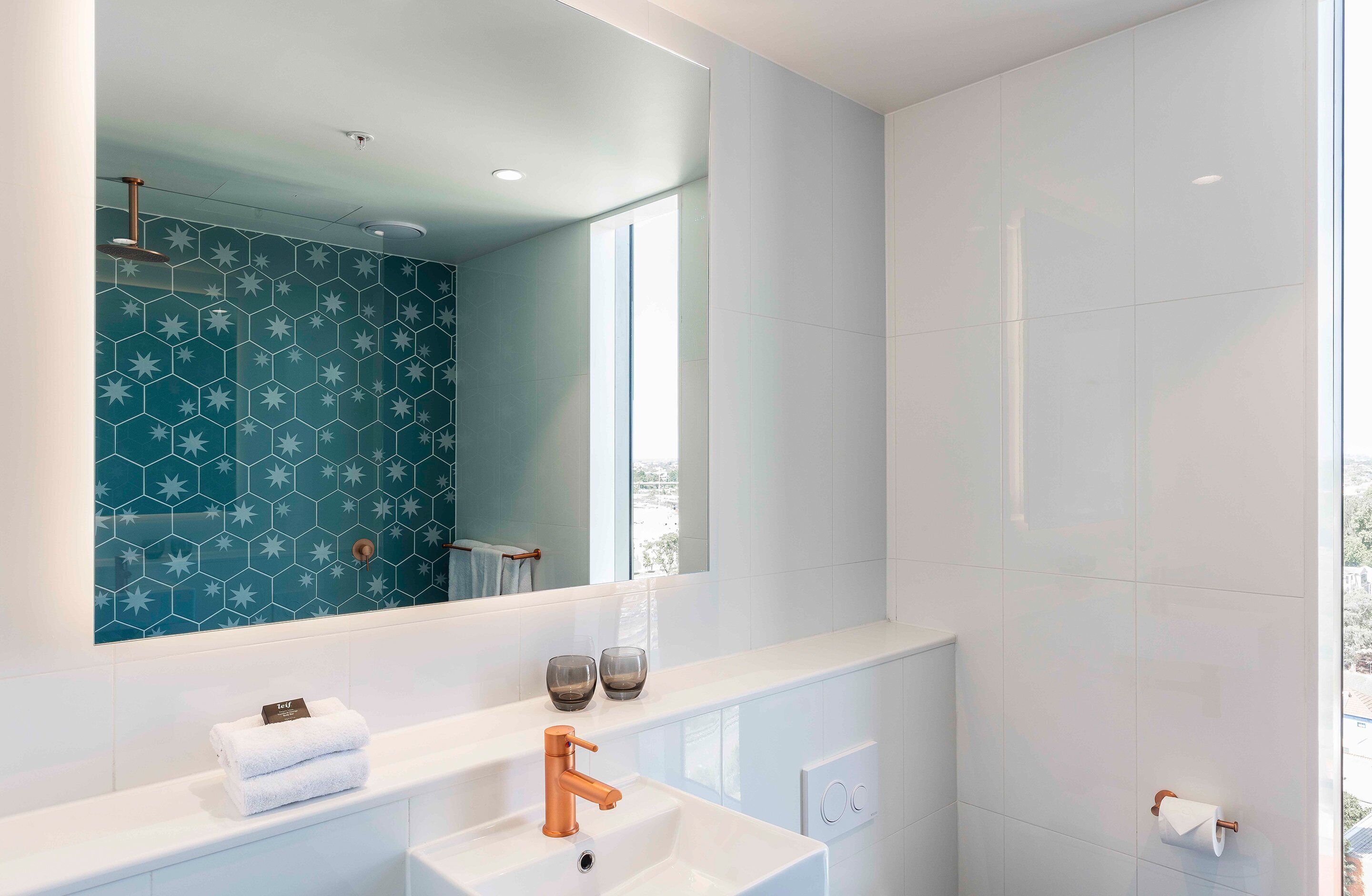 A modern bathroom with a large mirror, two sinks, a shower, and a window providing natural light.