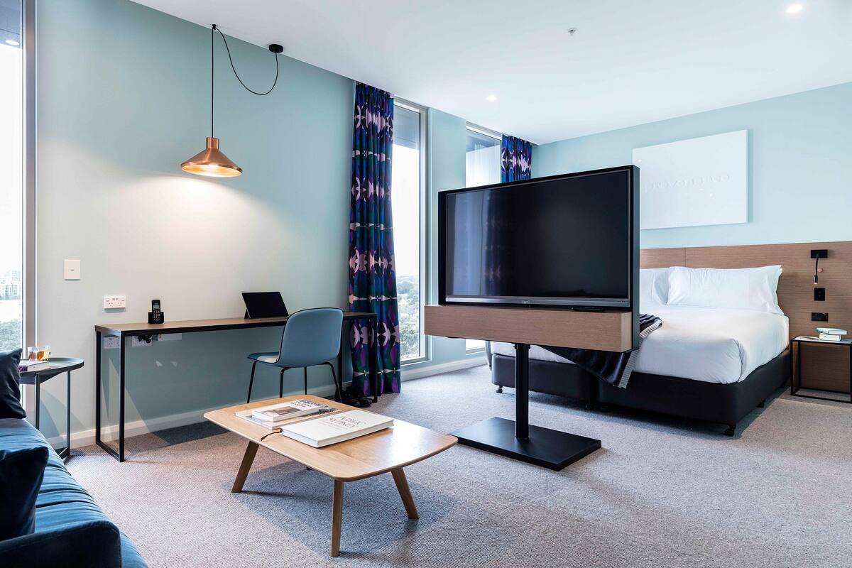 A modern hotel room with a large flat-screen TV, a bed with white linens, a desk with a laptop, and a coffee table with a book.