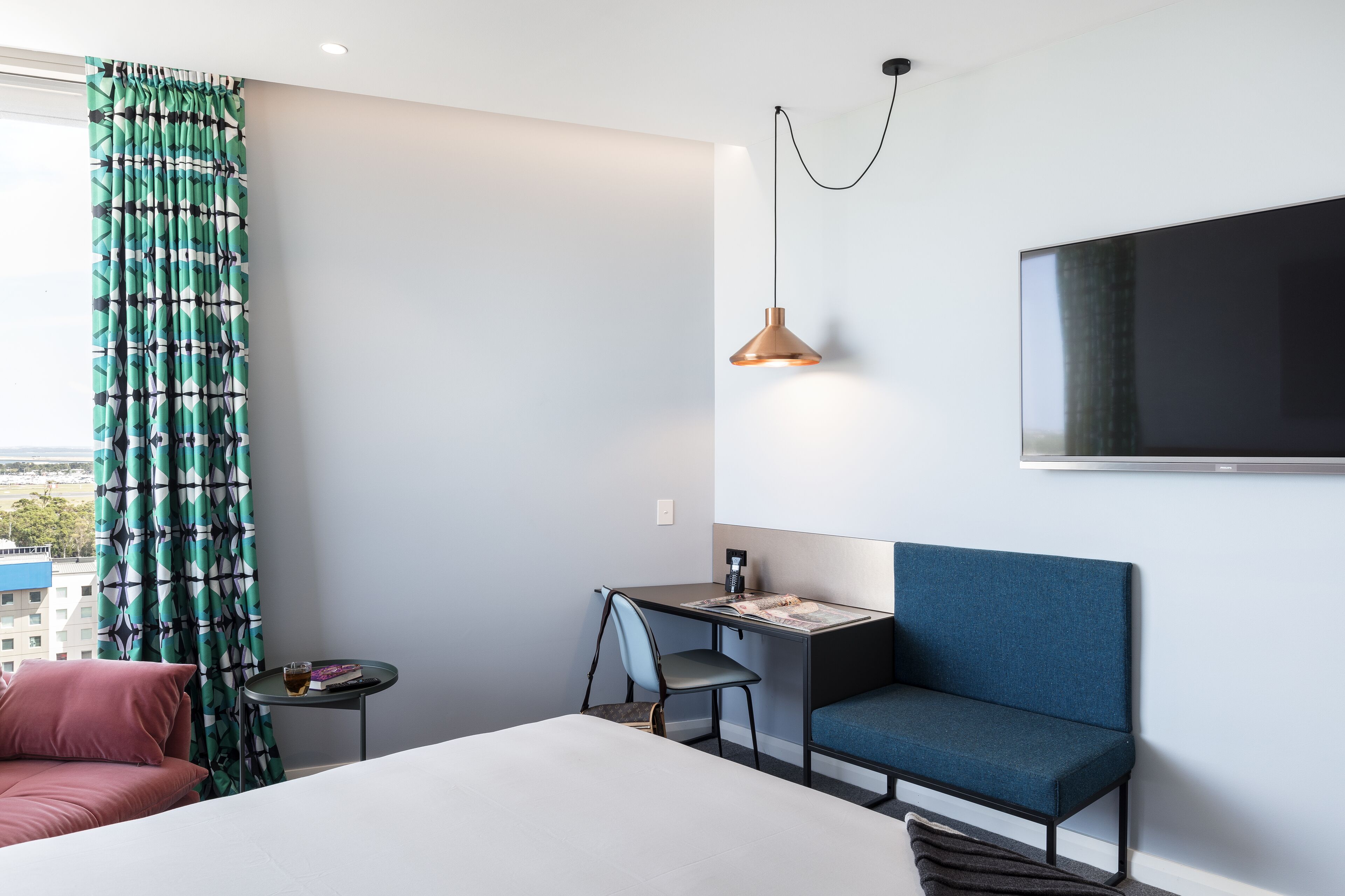 A modern hotel room with a bed, a desk, a chair, a TV, and a view of the city.