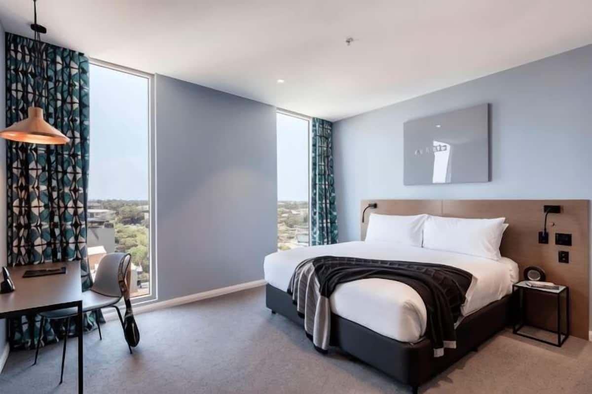 A modern hotel room with a large bed, a desk, a chair, a desk lamp, and a view of the city.
