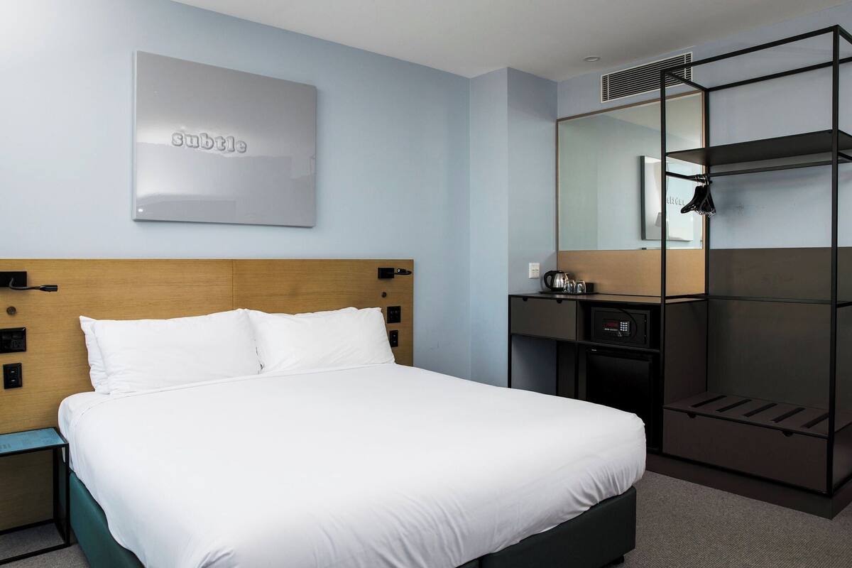 A modern hotel room with a neatly made bed, a wooden headboard, a built-in wardrobe, and a wall-mounted mirror.