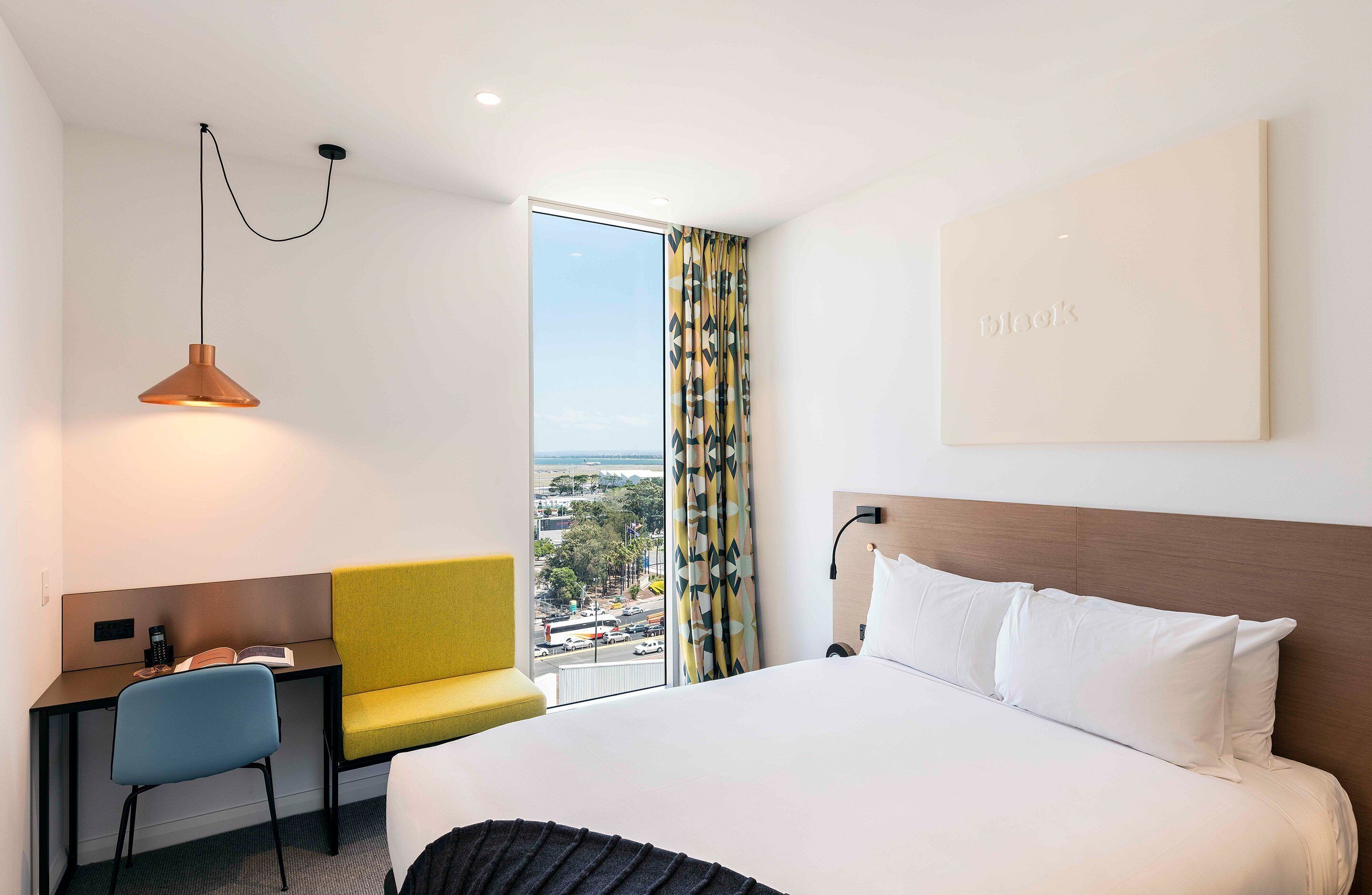 A hotel room with a bed, a desk, a chair, a window with curtains, and a view of the city.