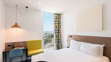 A hotel room with a bed, a desk, a chair, a window with curtains, and a view of the city.