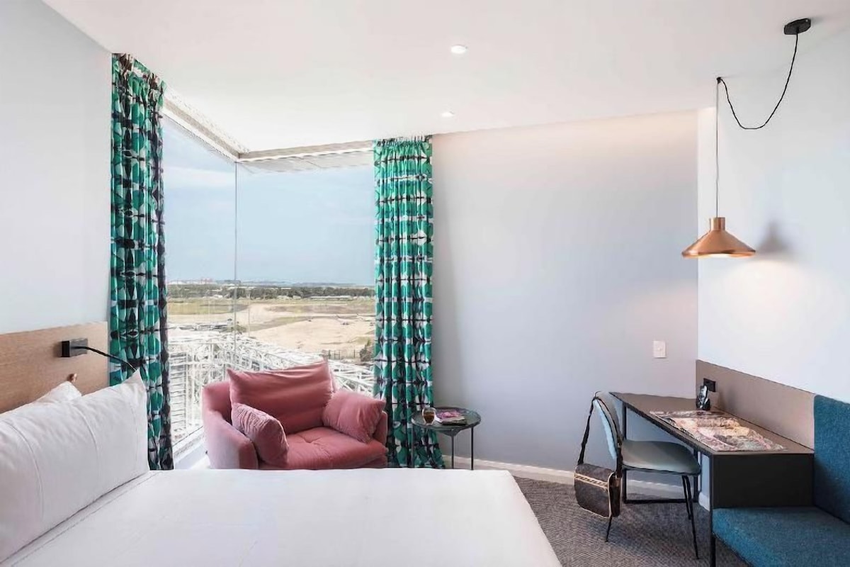 Una habitación de hotel con una cama, una silla rosa, una mesita pequeña, un sofá azul y vistas a un paisaje a través de amplios ventanales.