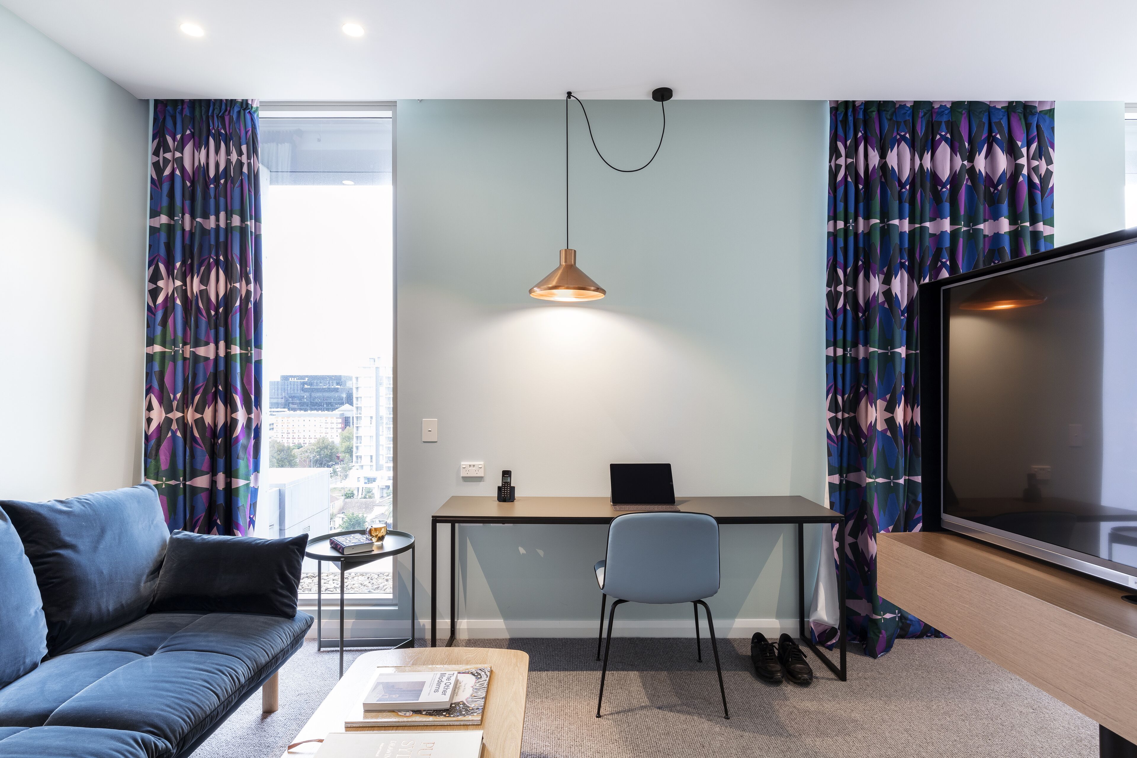 A modern living room with a sofa, a coffee table, a TV, a desk with a laptop, and a pendant light.