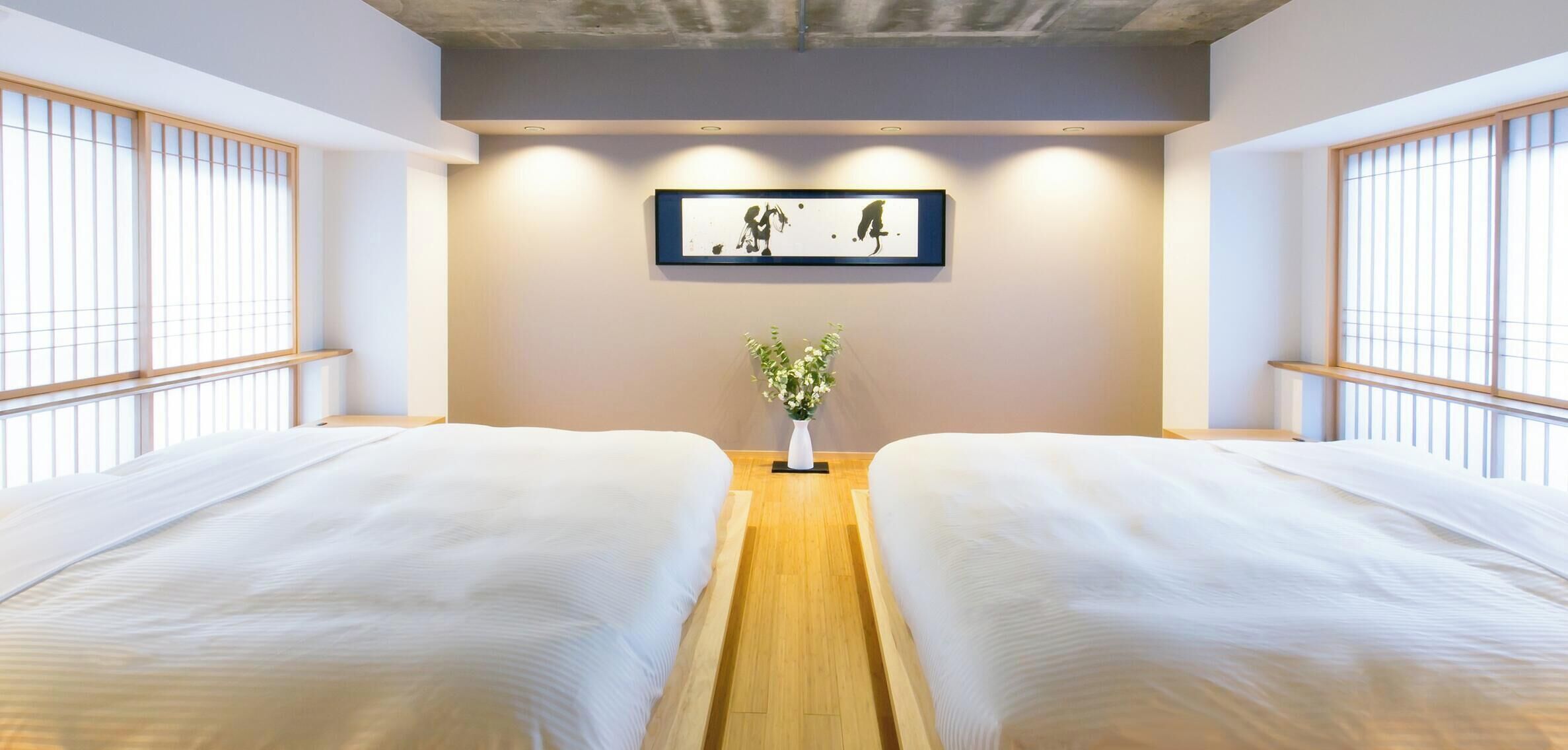A hotel room with two beds, a wooden floor, a framed artwork on the wall, and a potted plant.