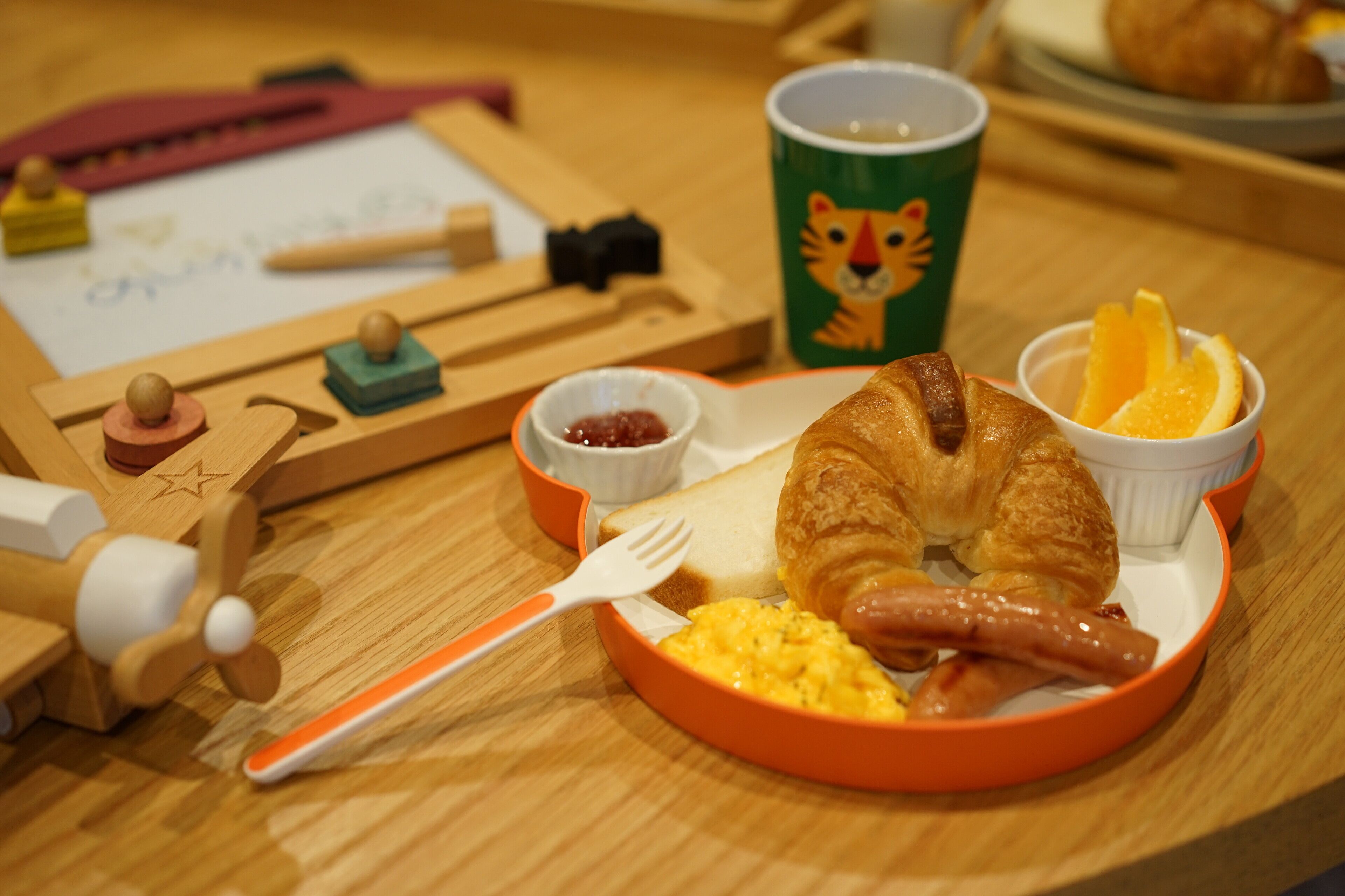 A breakfast setting with a croissant, scrambled eggs, sausage, orange slices, and a cup with a tiger design.