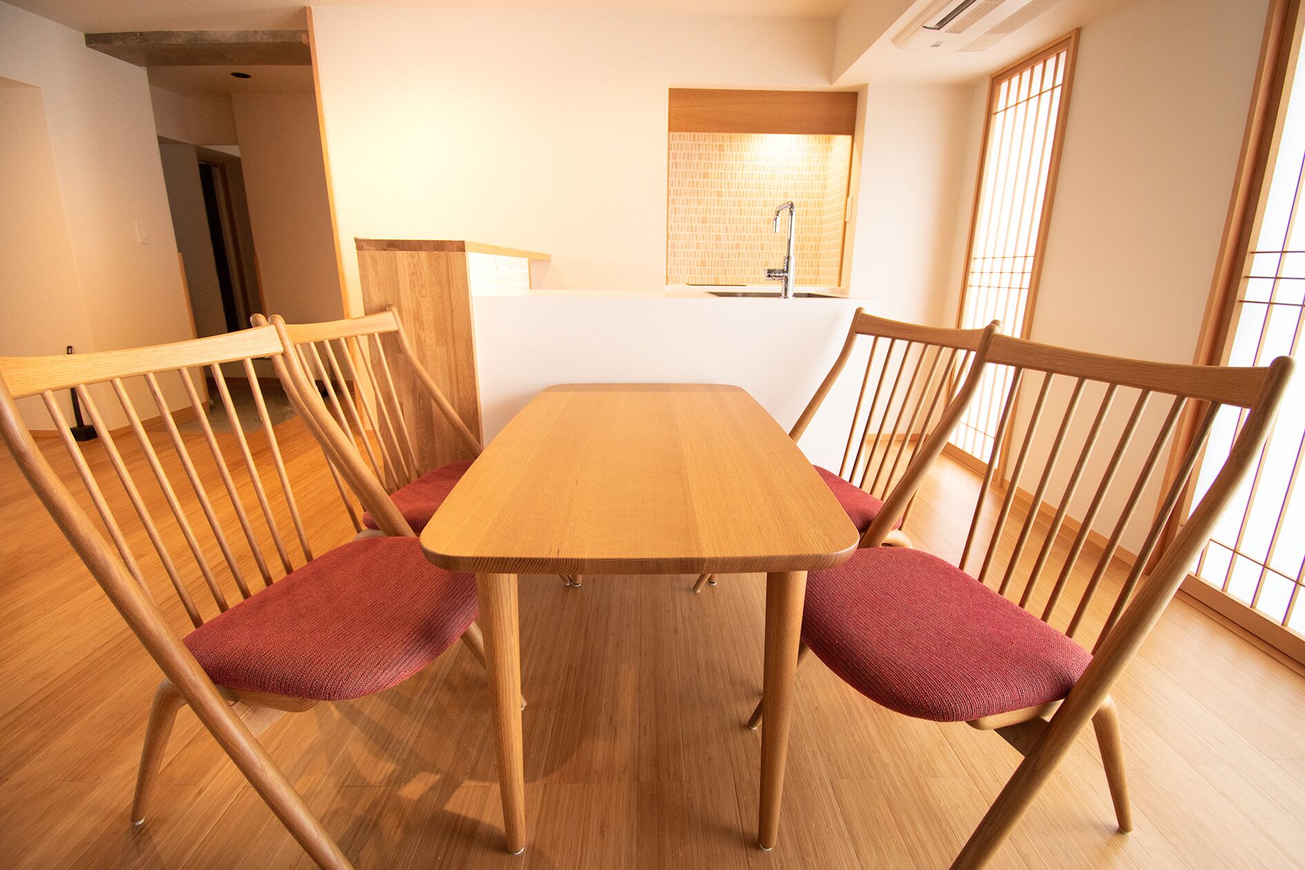 Una mesa de comedor de madera con cuatro sillas, cada una con un cojín rojo. El cuarto tiene piso de madera y una ventana con diseño de cuadrícula.