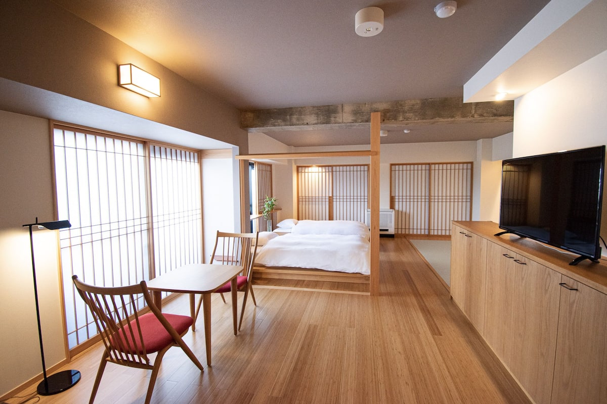 Een hotelkamer met een bed, een televisie, een bureau met stoel, en een raam met jaloezieën.