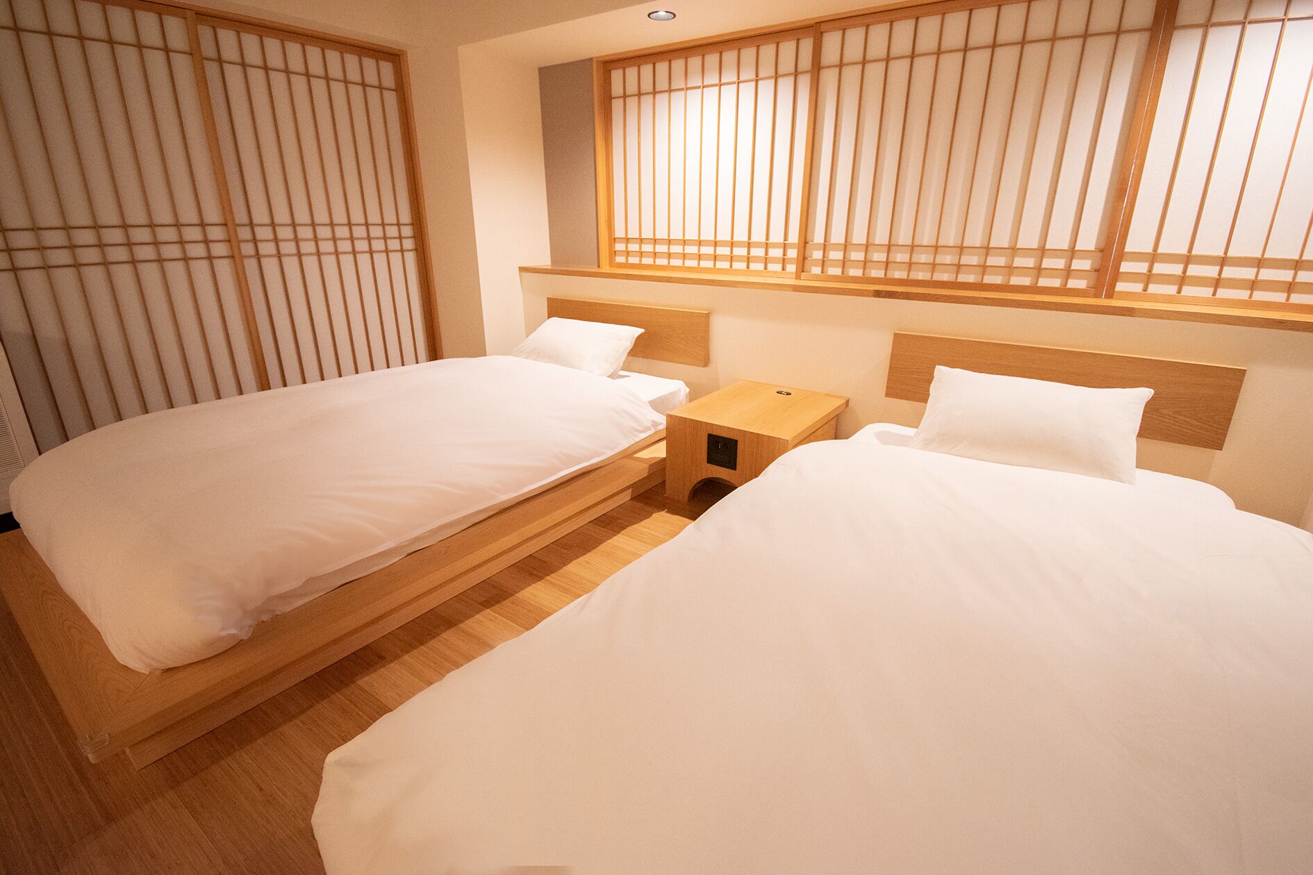 A hotel room with two single beds, wooden headboards, a small wooden nightstand, and a window with vertical blinds.