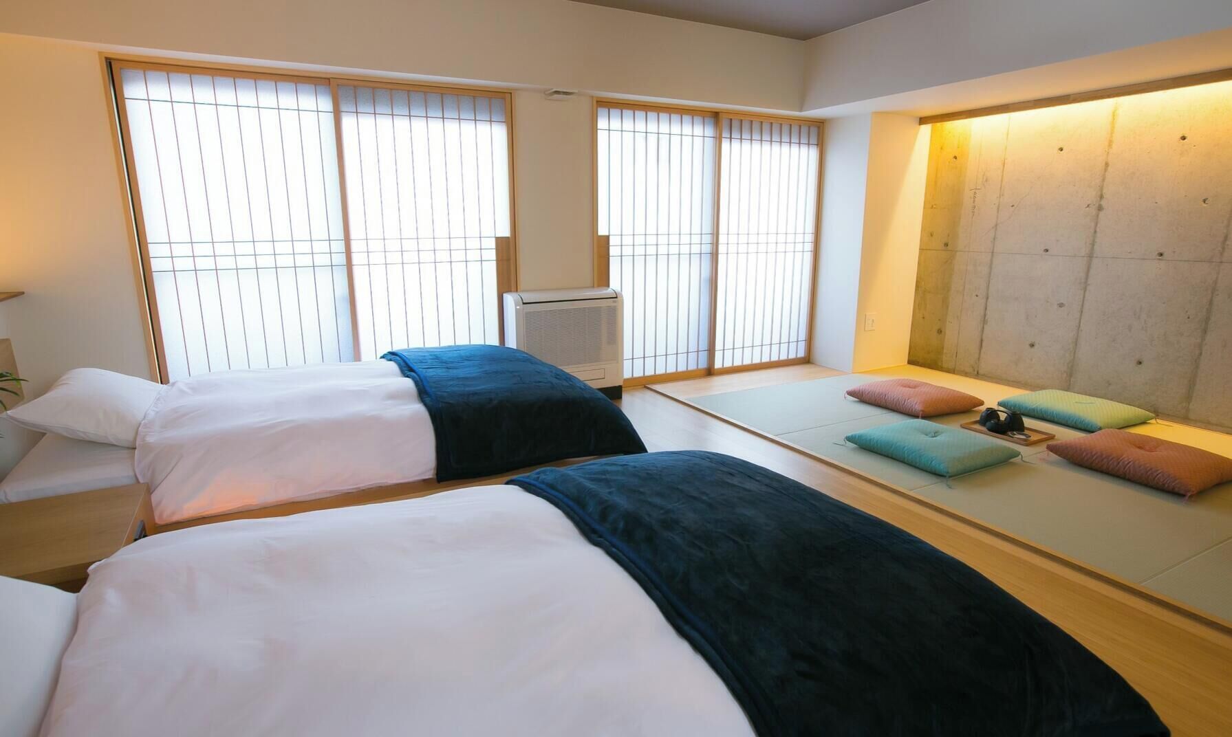 A hotel room with two beds, a futon, a heater, and large windows with blinds.