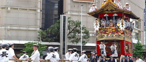A traditional Japanese parade with participants in white uniforms, a decorated float, and a building with a sign reading "LAOX Japan Quality Store".