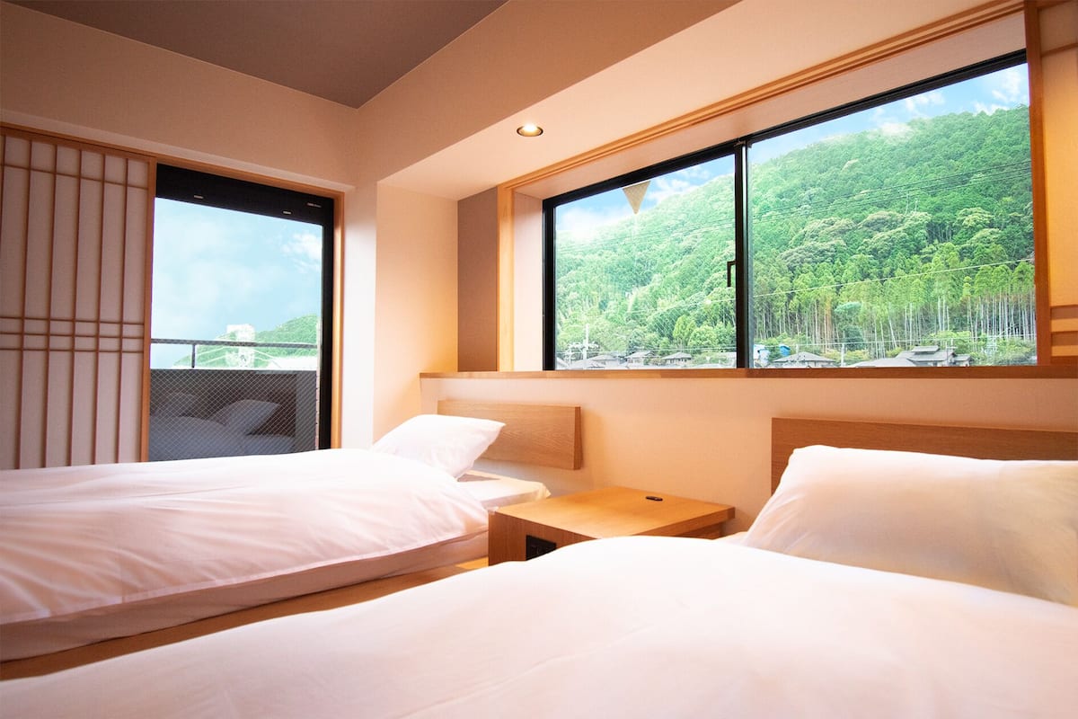 Een hotelkamer met twee bedden, een klein houten nachtkastje en een groot raam met uitzicht op een bebost gebied.
