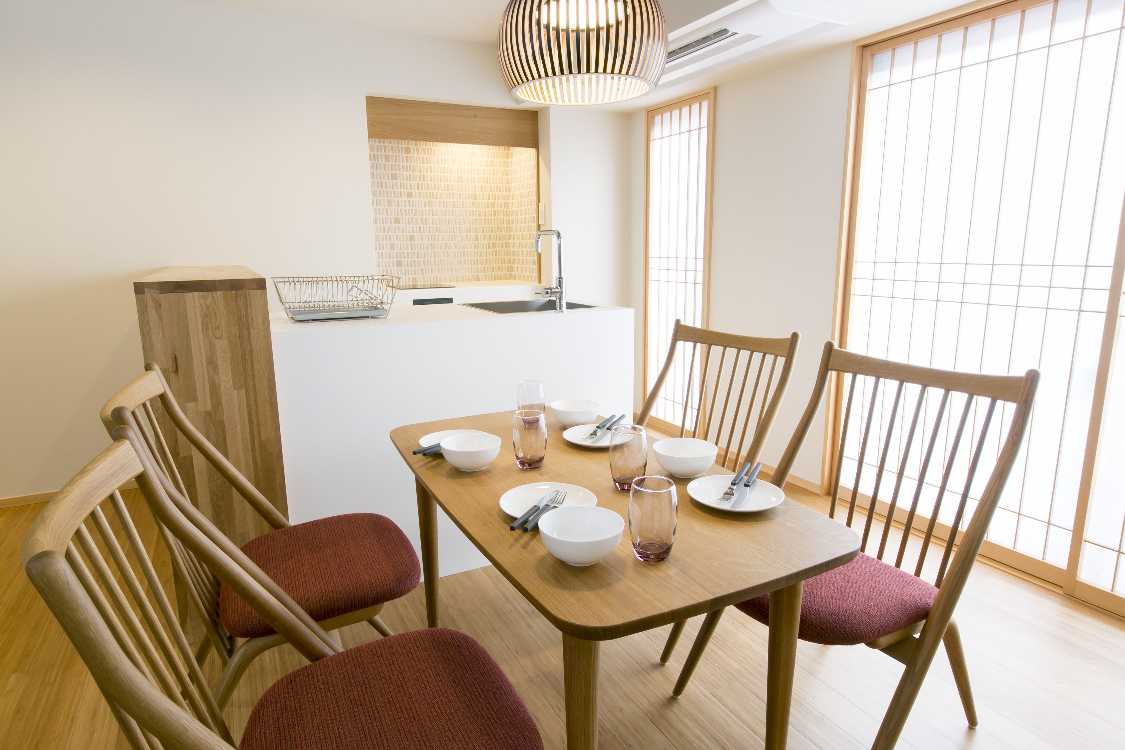 A dining area with a wooden table set for four, surrounded by wooden chairs with red cushions. A kitchen area is visible in the background with a tiled backsplash and a hanging light fixture.