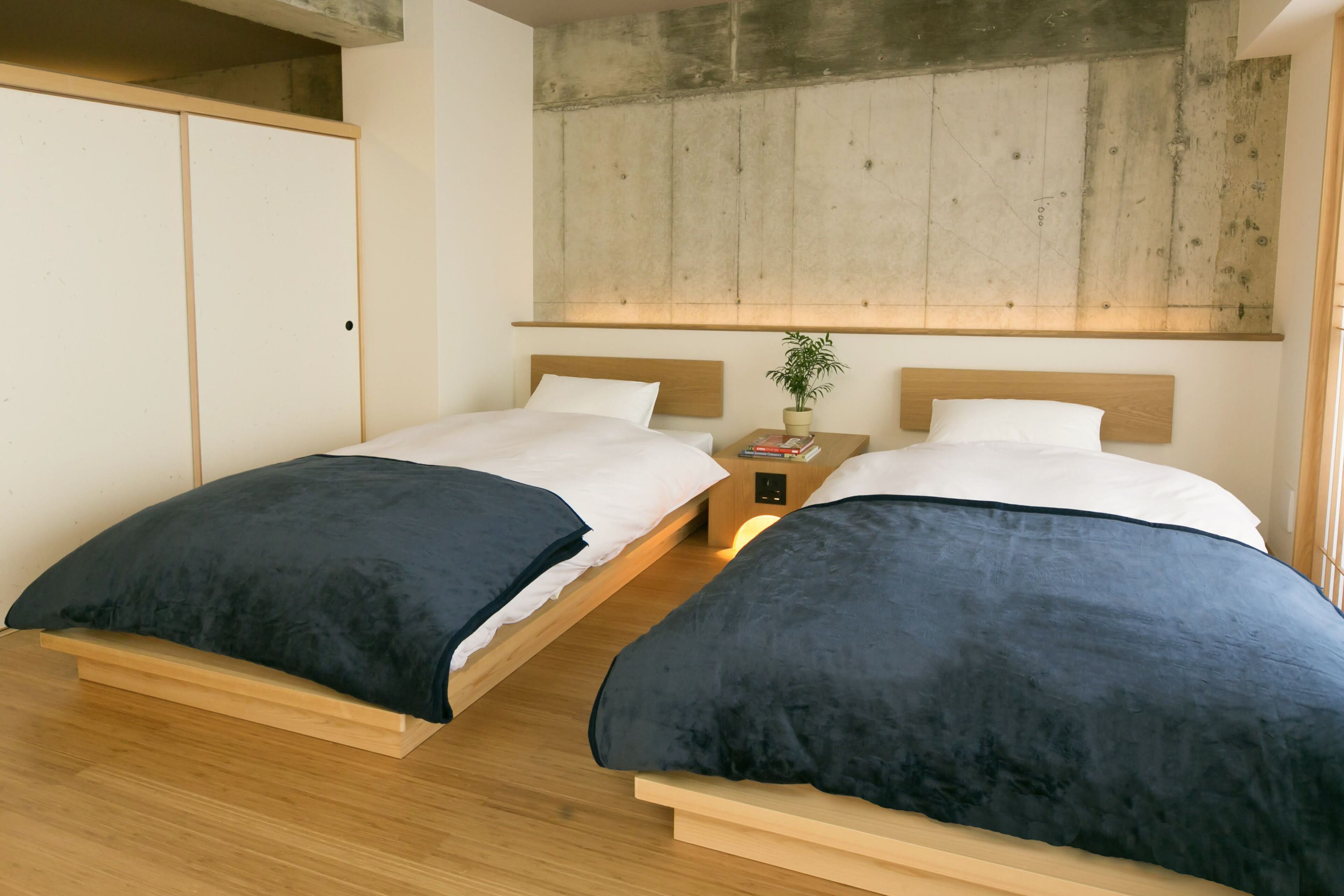 Habitación con cama doble, cabeceros de madera, una mesita de noche con una planta y pared de concreto.