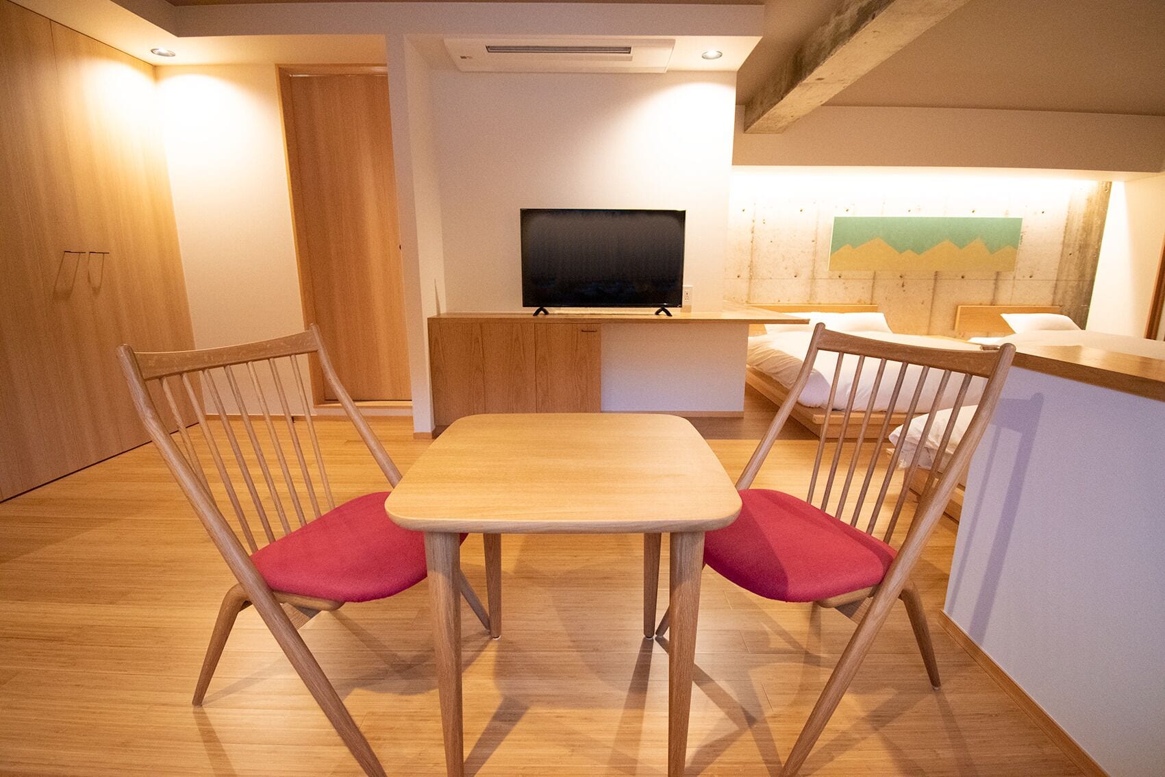 A compact living space with a wooden dining table and two chairs, a flat-screen TV on a stand, and a bed with a white headboard.