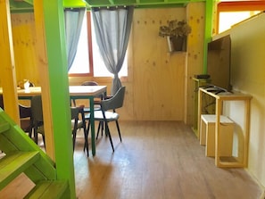 Una habitación con una mesa y sillas de madera, una ventana pequeña con cortinas y un televisor sobre un soporte de madera.