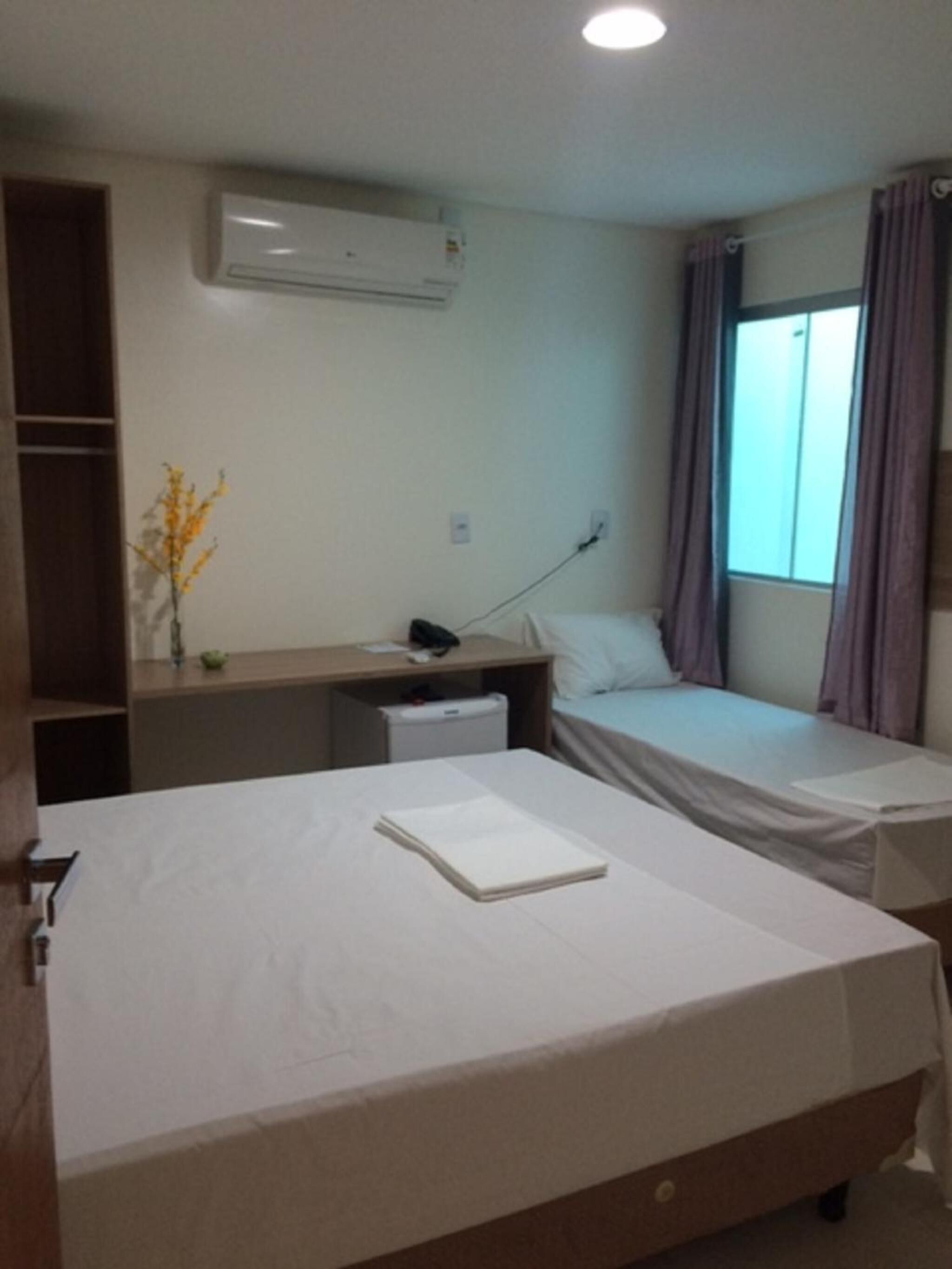 Habitación de hotel con dos camas individuales, un escritorio con silla, una mesita de noche, aire acondicionado en la pared, una ventana con cortinas y una planta en el escritorio.