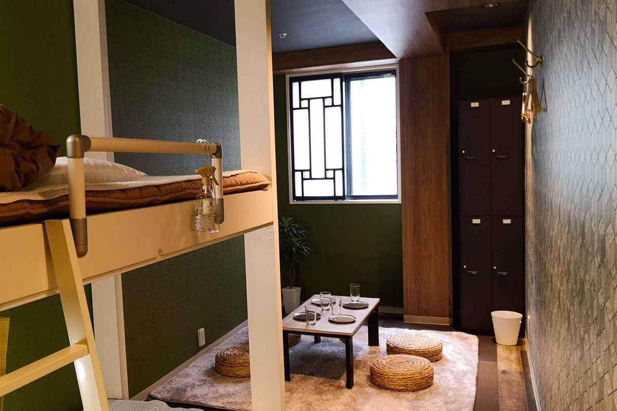 A bunk bed with a small table and chairs, a window with a view, and a wooden cabinet with drawers.