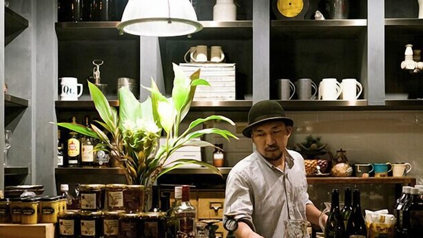 A chef in a kitchen with shelves stocked with jars, bottles, and kitchenware. - Atelier Café bar Seihei (Nakijin)
