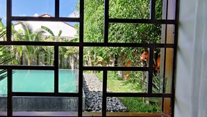 Una ventana con vistas a una piscina, vegetación y un edificio al fondo.