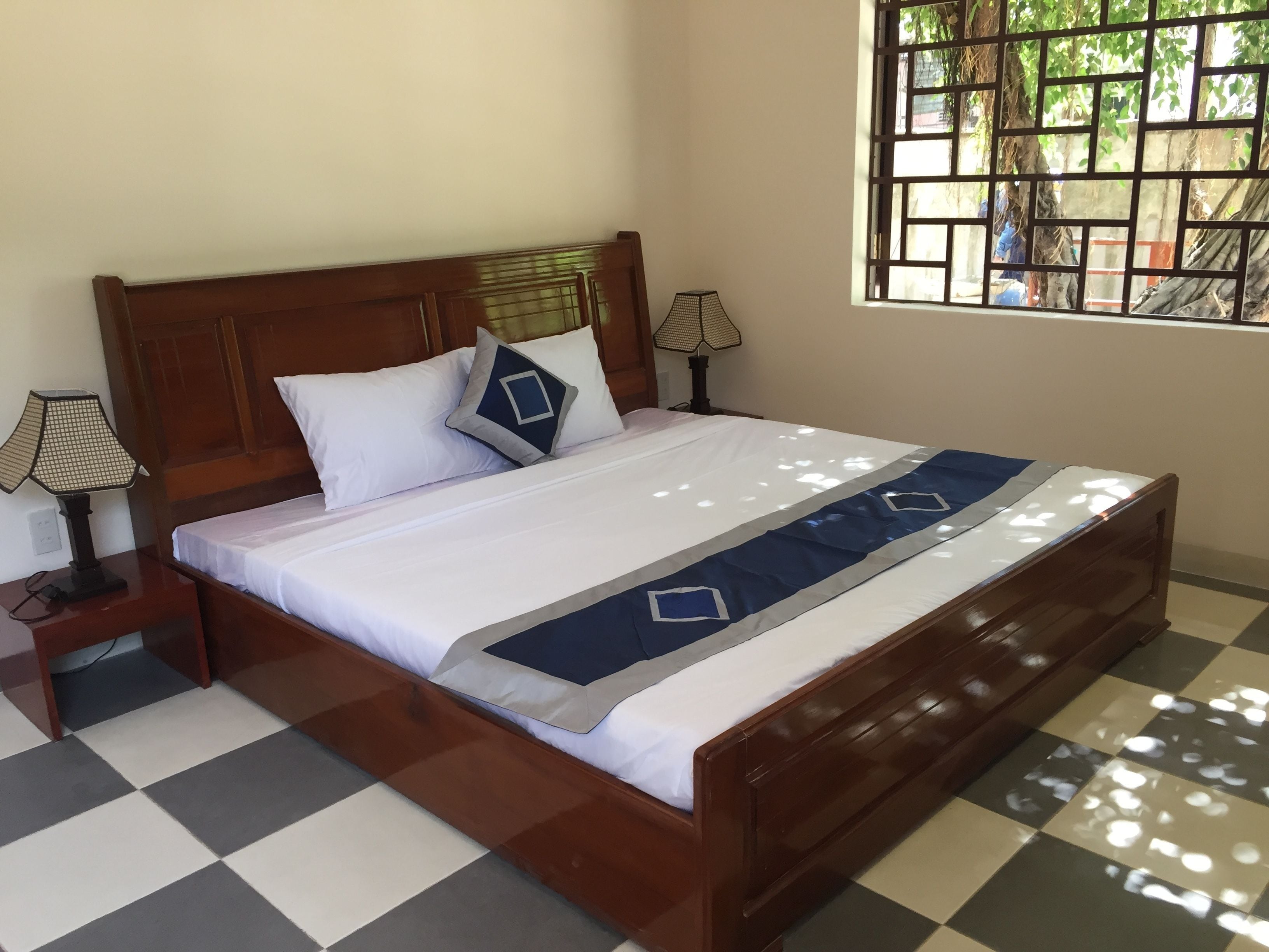 Una cama bien hecha con suelo a cuadros, un cabecero de madera y una ventana con vistas a árboles.