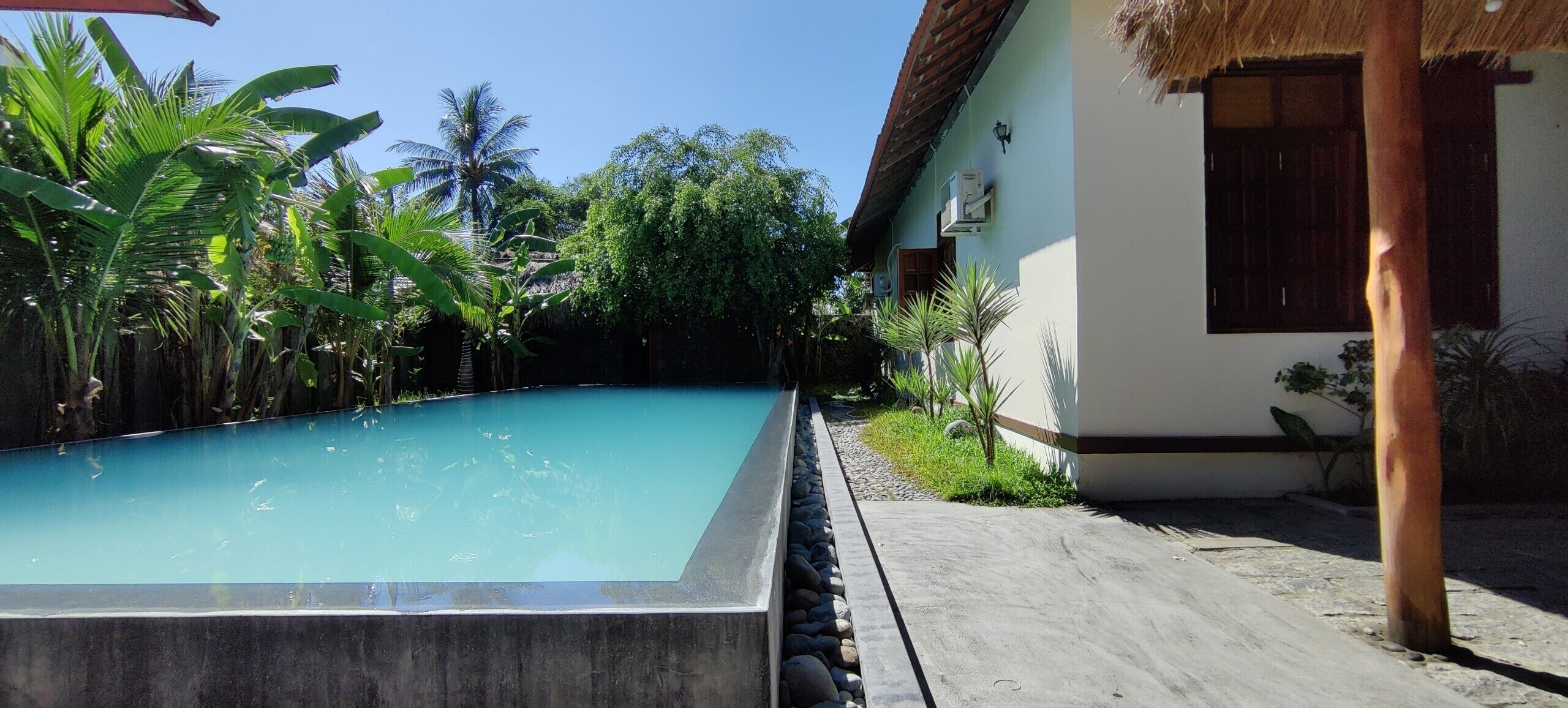 A rectangular swimming pool with a clear blue water surface, surrounded by lush greenery and a thatched-roof structure.