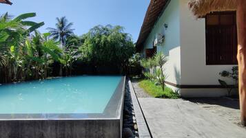 A rectangular swimming pool with a clear blue water surface, surrounded by lush greenery and a thatched-roof structure.
