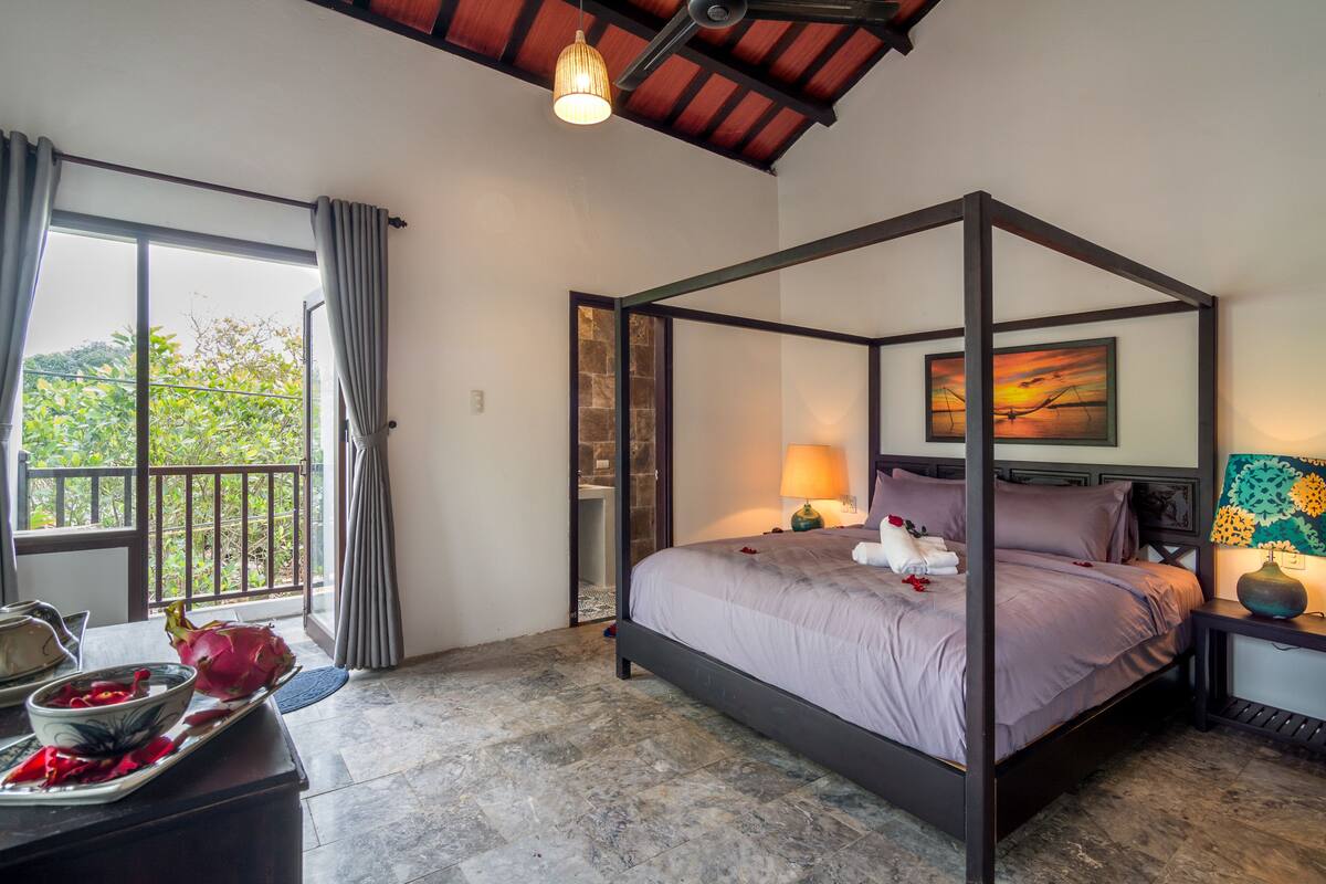 A four-poster bed with a view of greenery outside, a stone wall, and a sunset painting.