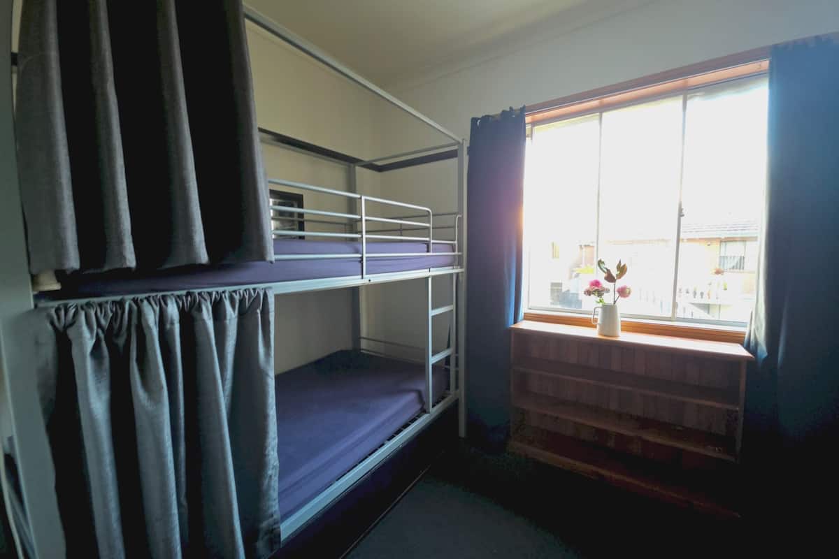 Une chambre avec un lit superposé, une fenêtre, des rideaux et une petite table sur laquelle est posé un vase.
