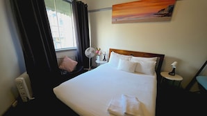 A hotel room with a bed, a window with curtains, a wall-mounted painting, a bedside table with a lamp, and a standing fan. - Coogee Beach House - Hostel (Coogee)