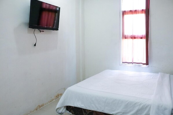 A small, sparsely furnished room with a single bed, a television mounted on the wall, and a window with curtains. - Graha Matahari (Surabaya)