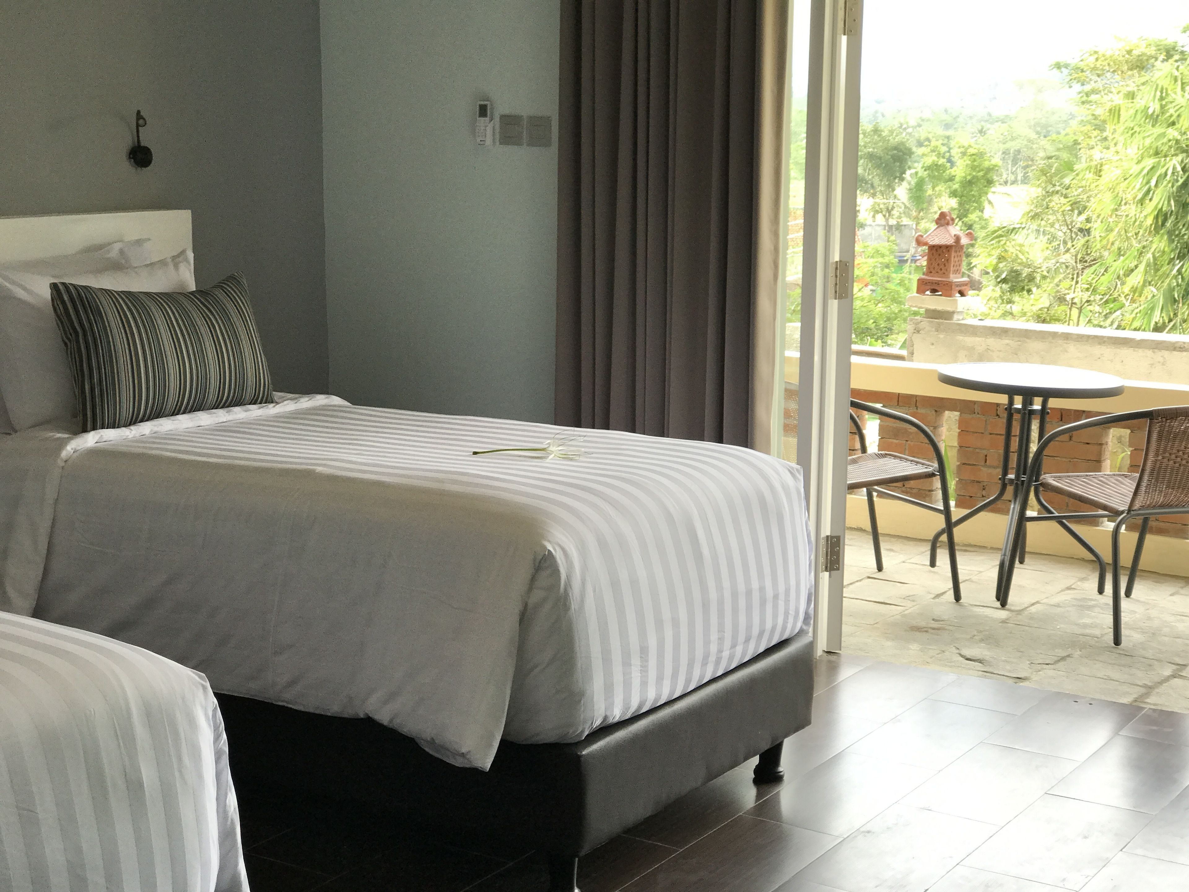 A hotel room with a bed, a small balcony with a table and chairs, and a view of greenery outside.