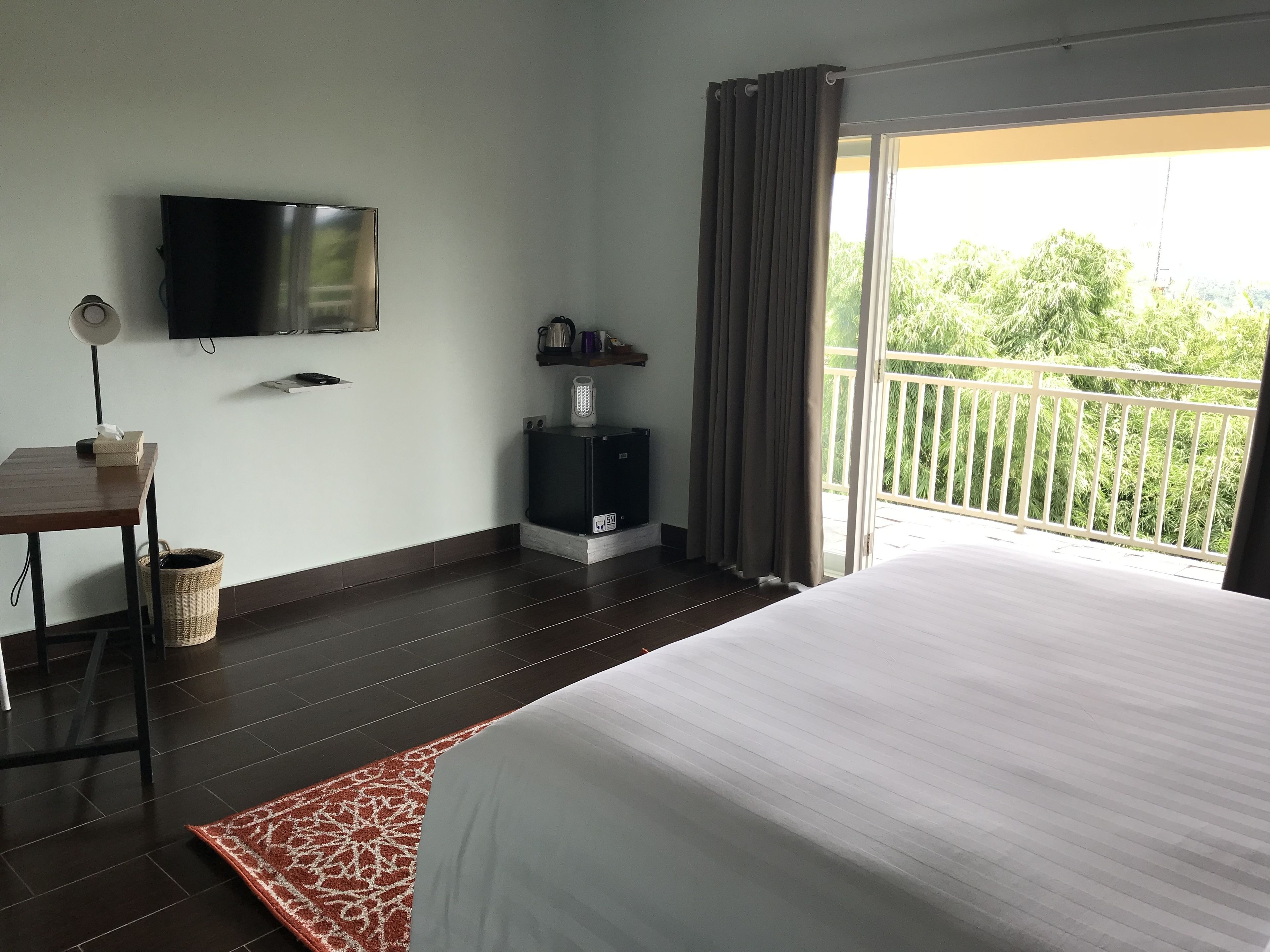 A hotel room with a bed, a TV mounted on the wall, a small table with a lamp, a waste basket, and a mini-fridge.