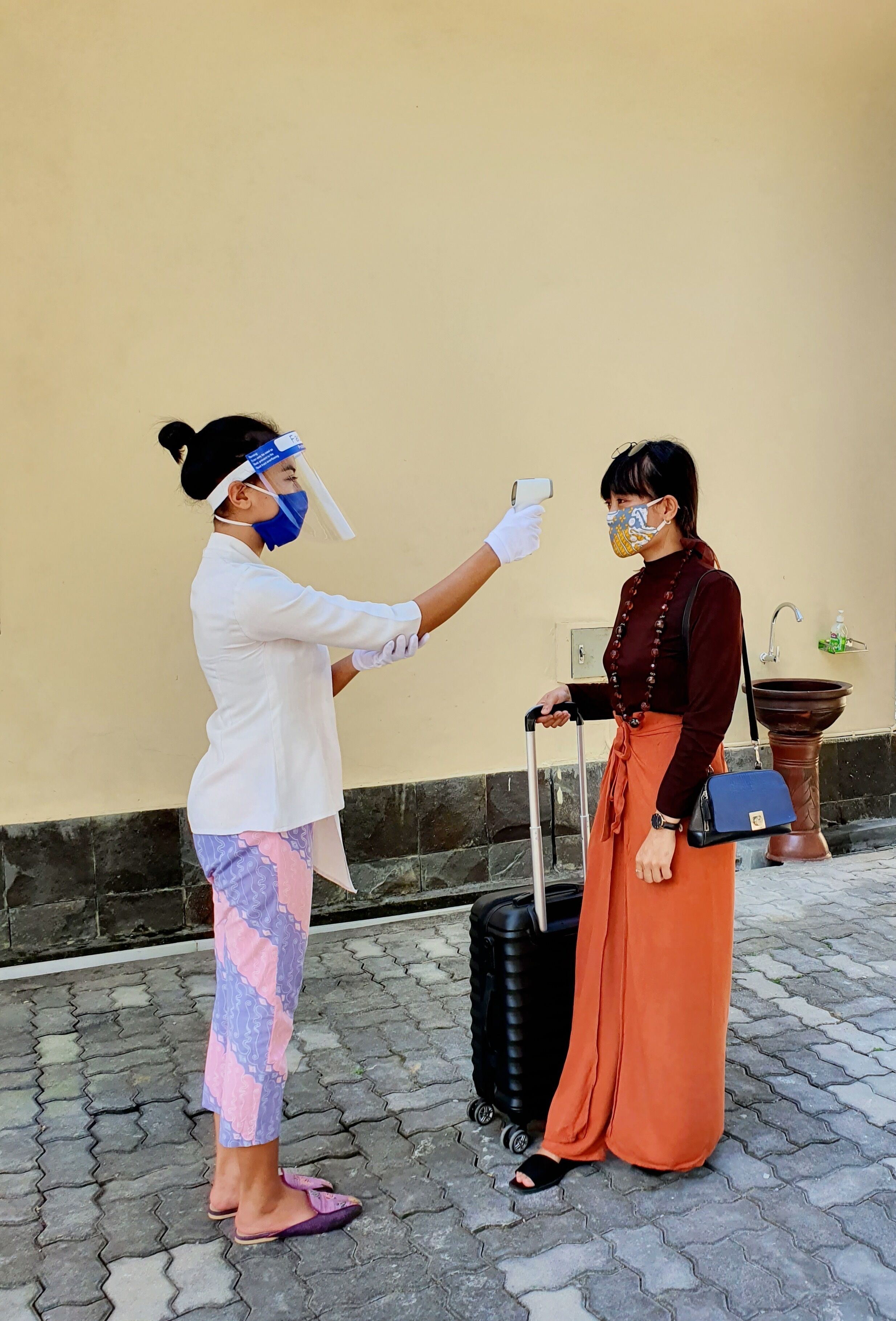 A person wearing a white shirt and colorful leggings standing next to another person with a suitcase, both wearing masks.