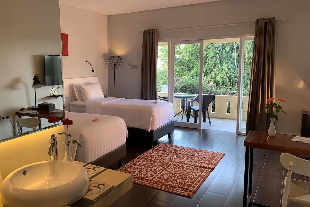 Une chambre d’hôtel dotée d’un grand lit, d’un coin salon avec une table et des chaises, d’une salle de bain avec un lavabo et un miroir, et d’un balcon offrant une vue sur la verdure.