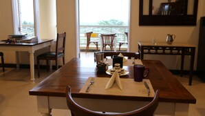 Un comedor con una mesa de madera dispuesta para dos comensales, una mesita auxiliar con una jarra y una taza, y vistas al balcón a través del ventanal.