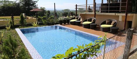 A rectangular swimming pool with a tiled edge, surrounded by a chain-link fence. There are wicker chairs with cushions and a wooden deck area.
