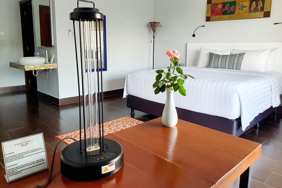 Une chambre d’hôtel comprenant un lit, une table de chevet, un vase avec des fleurs, une lampe de sol et un distributeur d’eau.