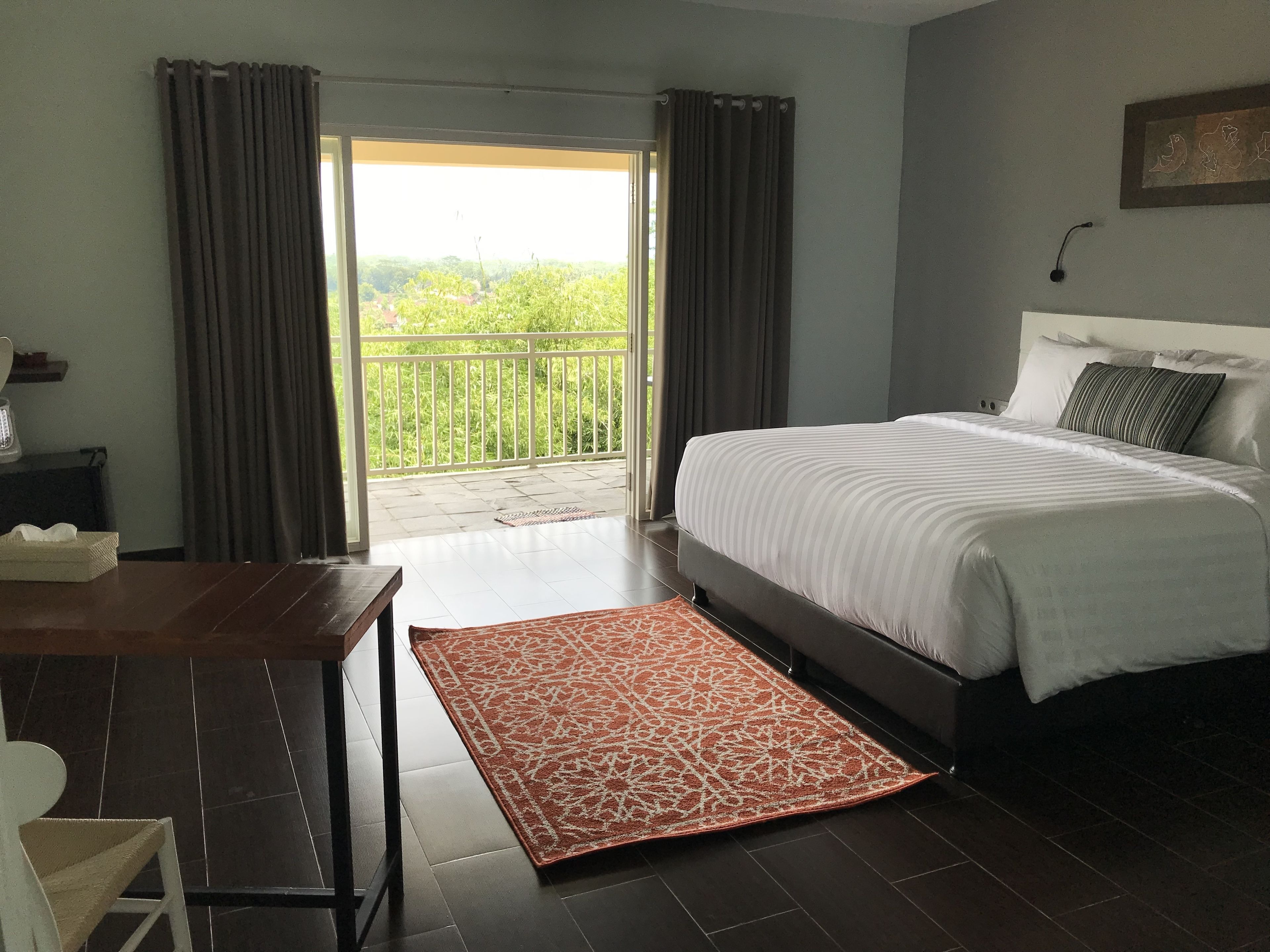 A bedroom with a bed, a desk, a chair, a patterned rug, and a balcony with curtains.